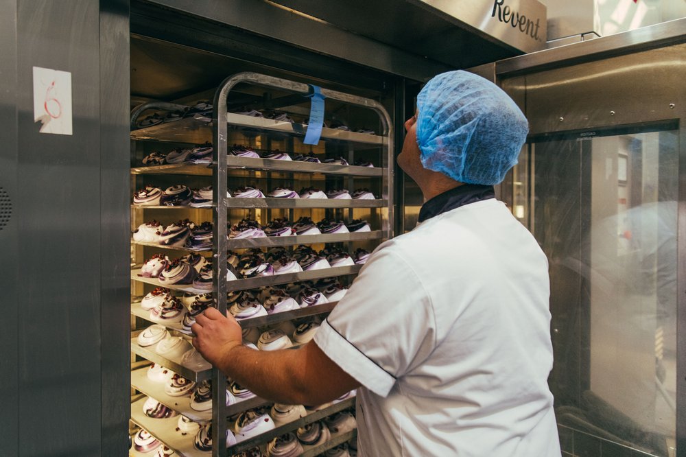 A man in a blue hair net and a white chef's jacket wheels trays of meringues into a large fridge