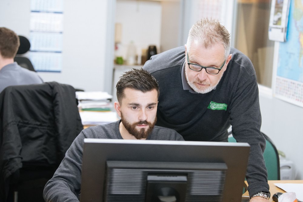 An older man in a grey sweatshirt looks over the shoulder of a younger man working at a computer