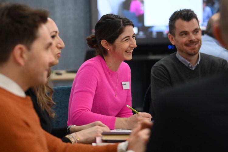 A woman in a bright pink jumper sits amongst three other people who are all slightly out of focus. They are all looking towards a speaker who is off-camera