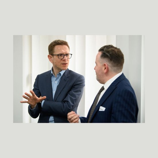Anthony Impey, dressed in a blue suit and a blue check shirt, speaks to a man in a pinstripe suit who is turned away from the camera