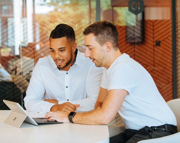 Business leader and employee in white shirts sitting at white table in modern office with glass walls looking at business finances on smart tablet and keyboard