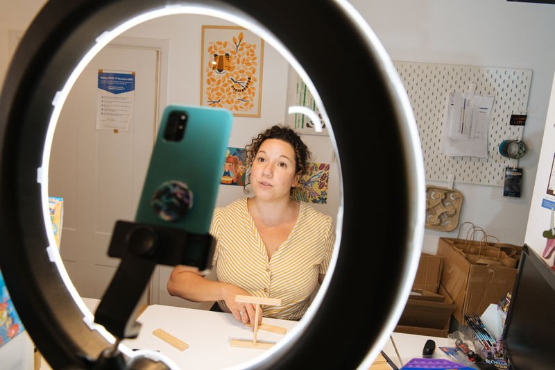 A woman in a yellow blouse films video content in front of a desk light