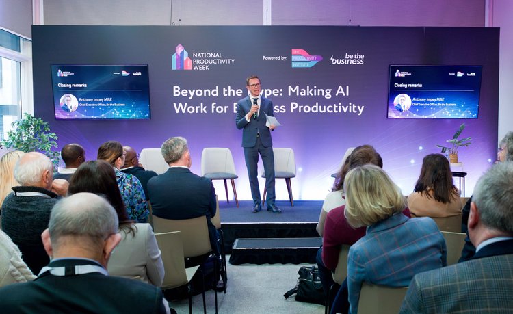 Anthony Impey stands in front of a purple screen with the words 'Beyond the Hype: Making AI Work for Business Productivity'. He is speaking to an audience who have their backs to the camera